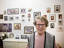 Seniorin steht in ihrem Zimmer vor einer Wand mit Familienfotos