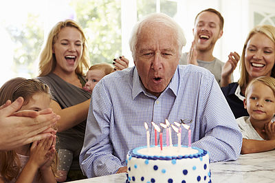 Senior pustet im Kreis seiner Familie die Kerzen einer Geburtstagstorte aus.