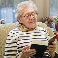 Seniorin sitzt in einem Sessel und ließt ein Buch.